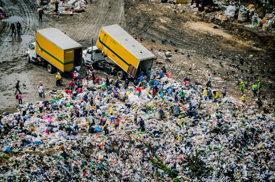 An aerial view of a large open waste site filled with numerous piles of mixed rubbish, including plastic bags, discarded packaging, and miscellaneous debris. Several individuals are seen actively coll